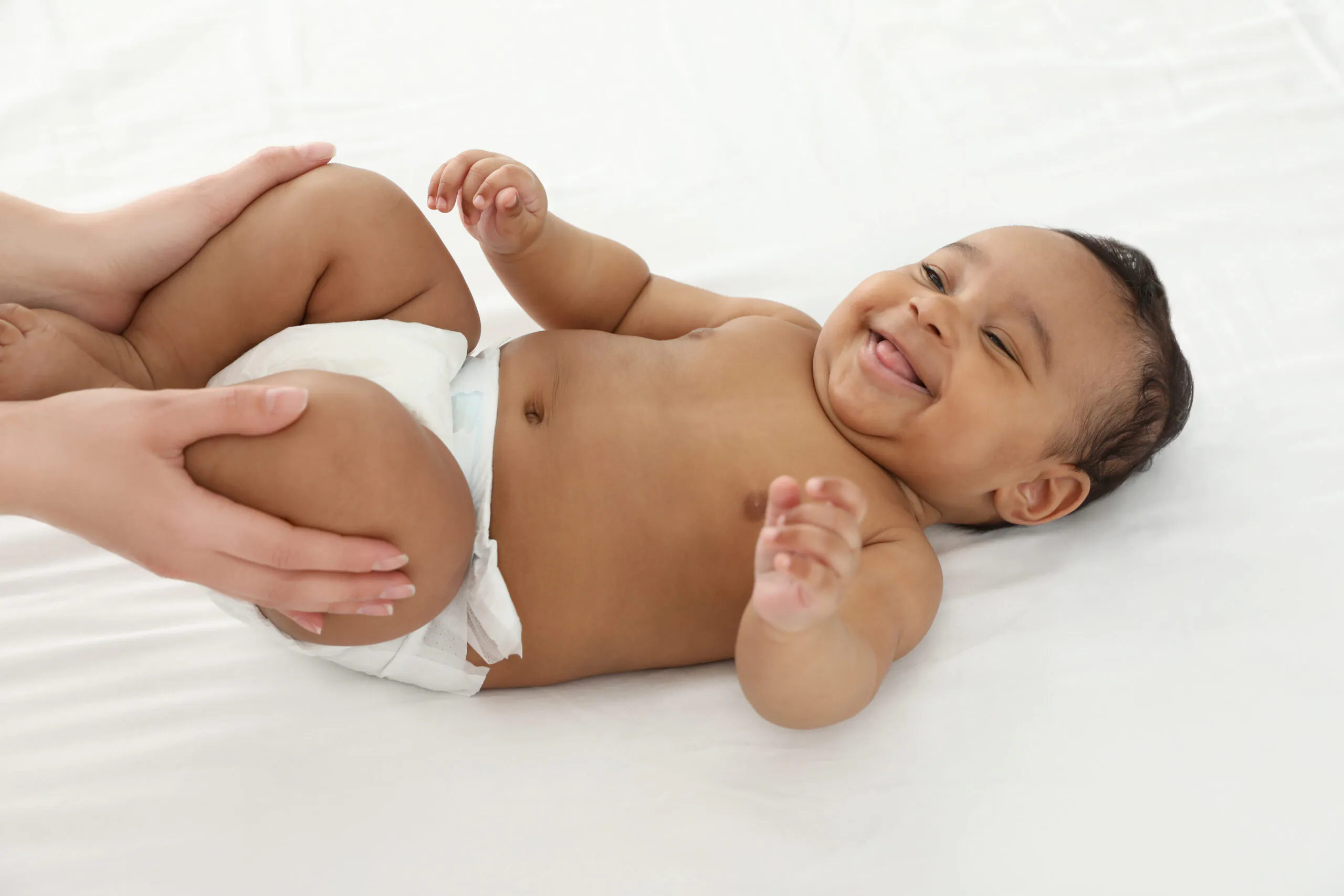 Un baby qui sourit et sa mère qui le masse sur un lit blanc pendant notre atelier massage bébé.